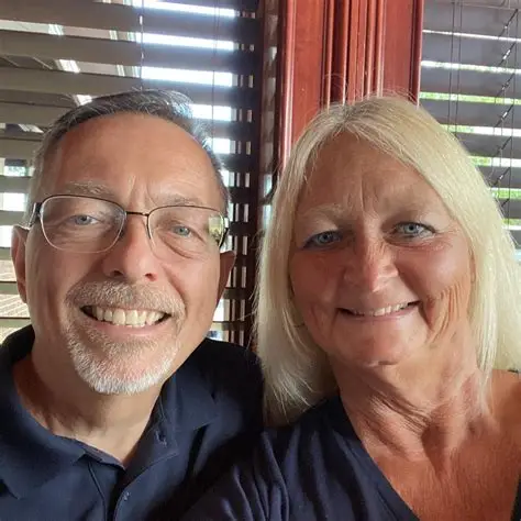 A smiling man with glasses and a blonde woman posing together indoors near window blinds.