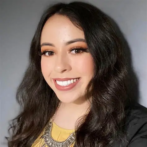Smiling woman with long dark wavy hair, wearing a yellow top and chunky silver necklace.