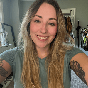 Smiling woman with long blonde hair and arm tattoos wearing a green t-shirt.
